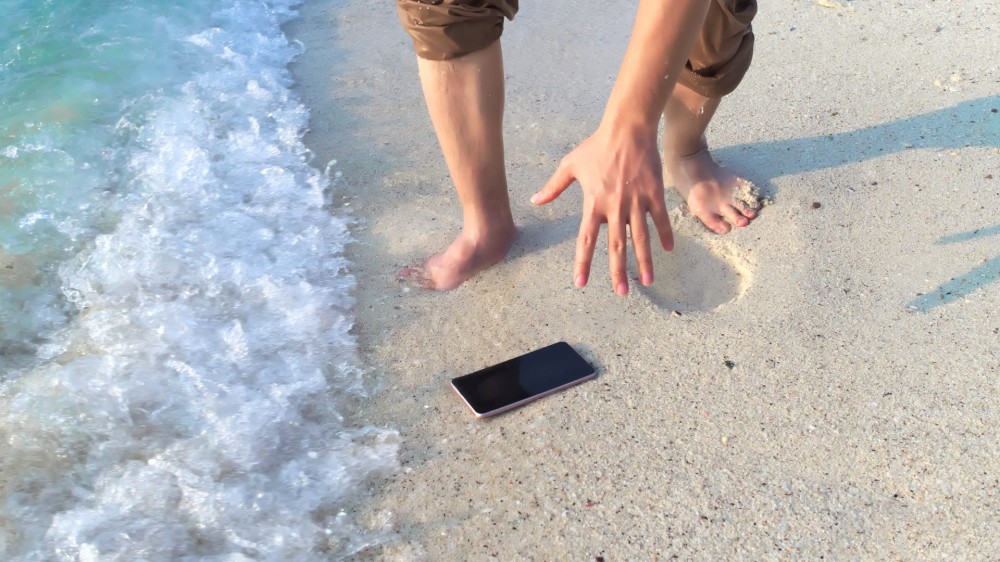 A man reaches for his dropped mobile smartphone on a tropical sandy beach, hoping to grab it before the waves wash over it.