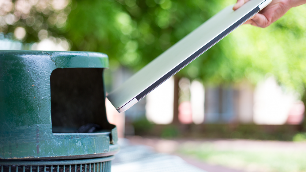 Closeup cropped portrait of someone tossing old notebook computer in trash can, isolated outdoors green trees background