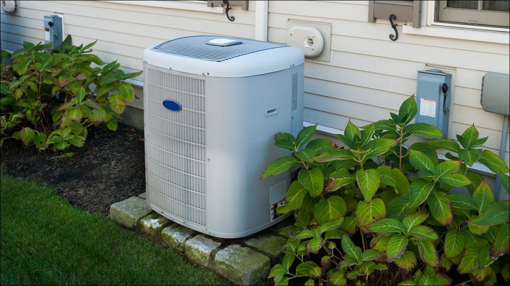 An air conditioner unit, sitting outside a home next to flowering bushes.